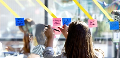 Una signora posiziona un foglietto Post-it® Super Sticky, 76 mm x 76 mm, su una parete di vetro per condividere l'acquisizione dei risultati delle attività con altri team o progetti