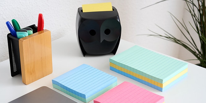 Una scrivania con Foglietti Post-it® di vari colori, un portapenne e un distributore di Foglietti Post-it® nero a forma di gufo
